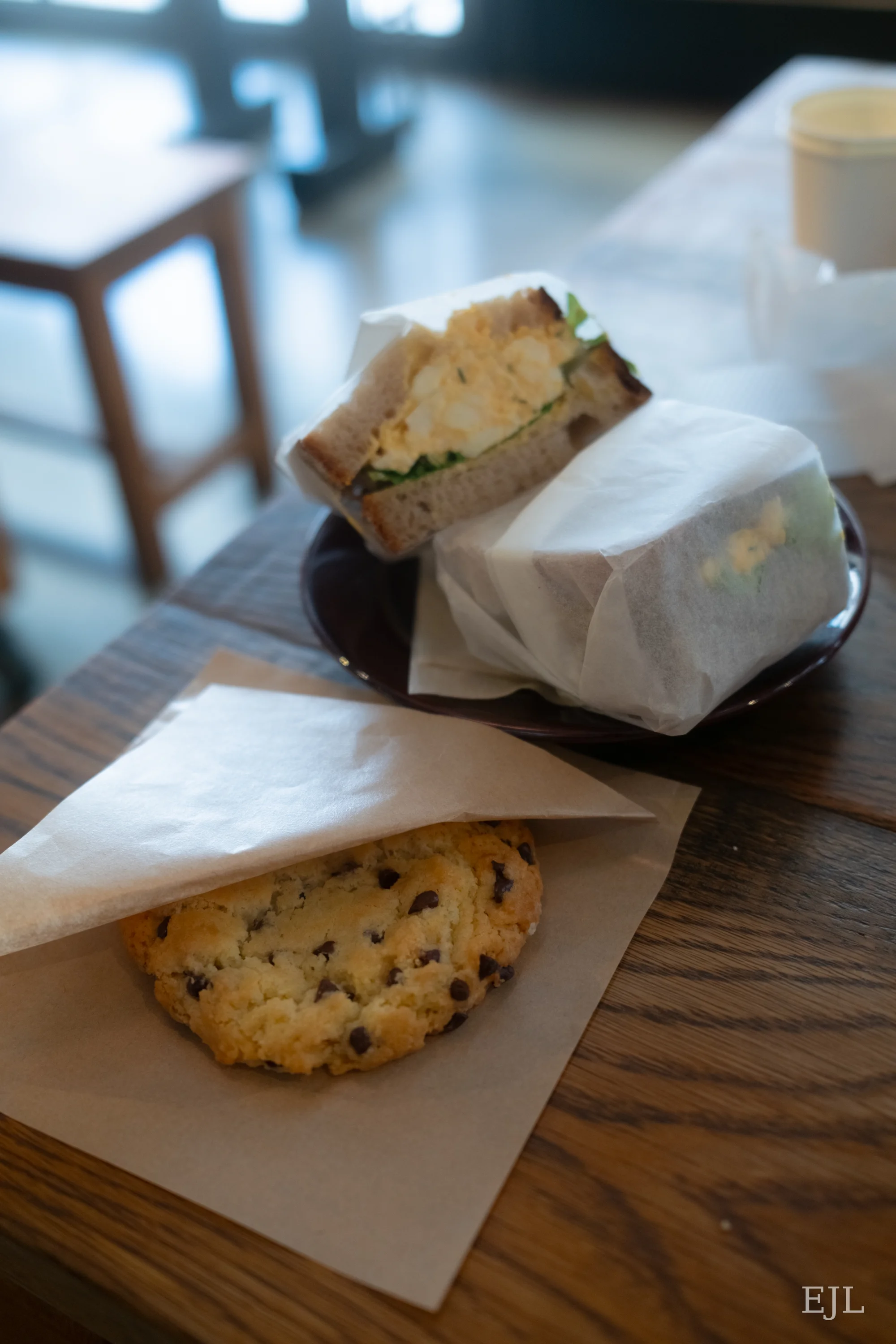 An egg sandwich in paper wrapping beside a chocolate-chip cookie on a cafe table