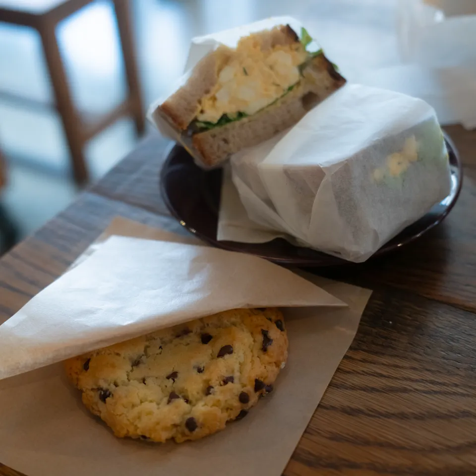 An egg sandwich in paper wrapping beside a chocolate-chip cookie on a cafe table