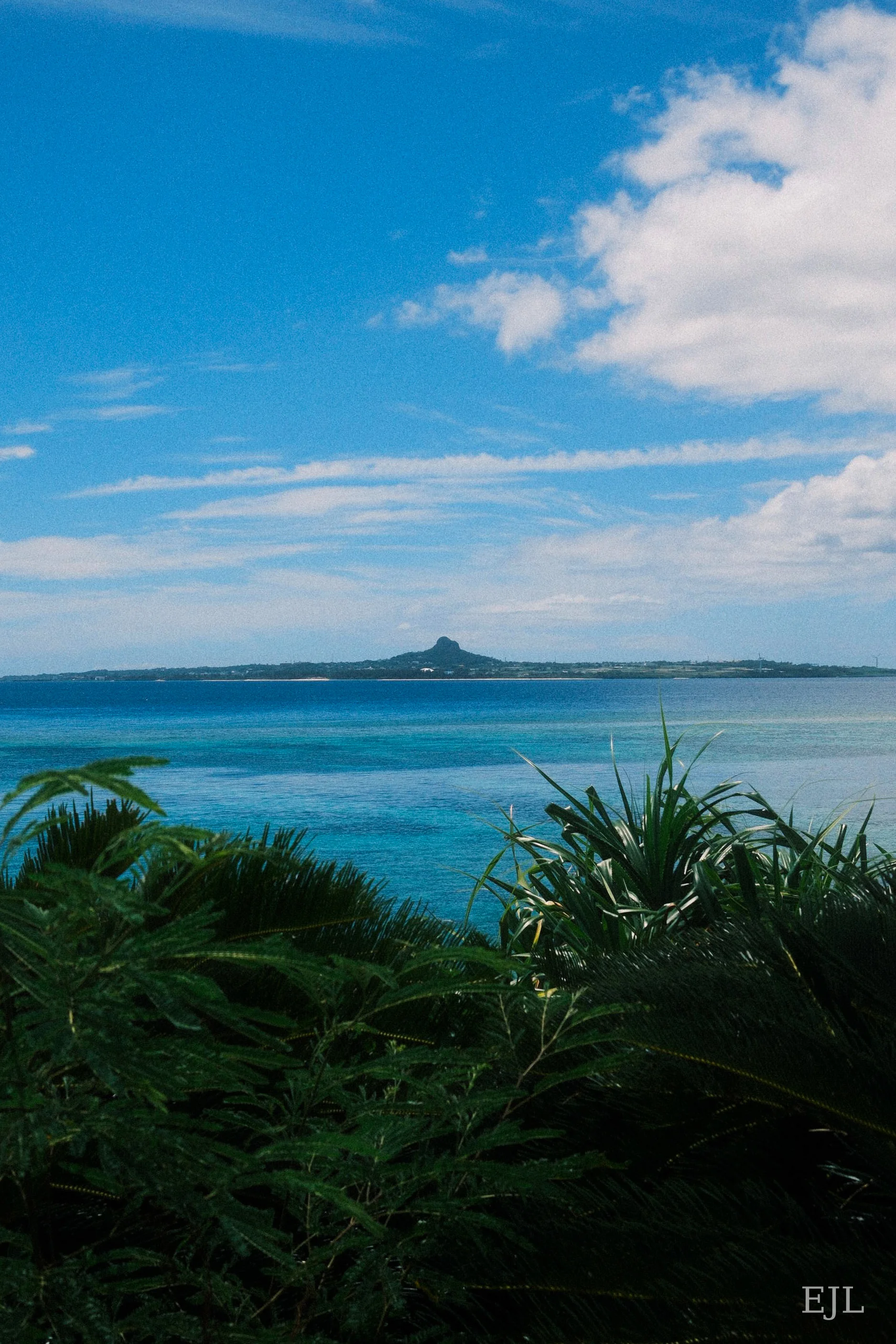 A distant island beyond turquoise sea and coastal greenery beneath a cloud-streaked sky