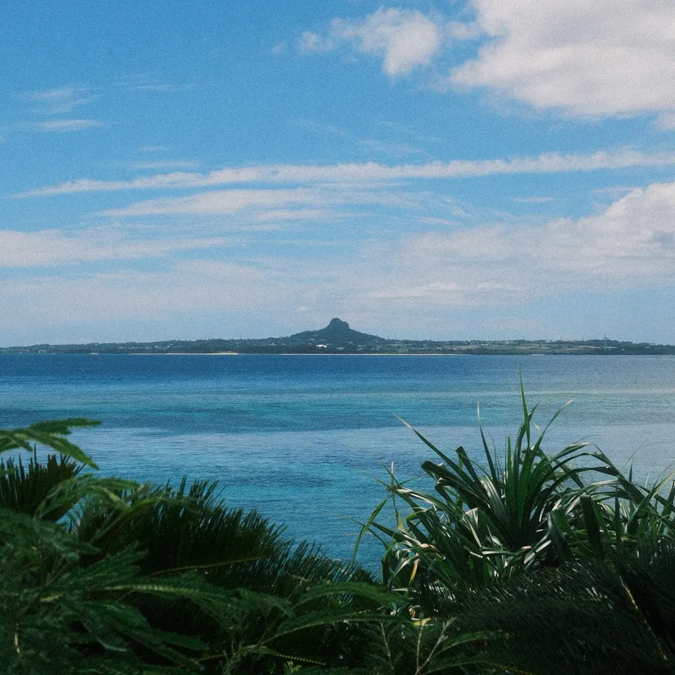 A distant island beyond turquoise sea and coastal greenery beneath a cloud-streaked sky