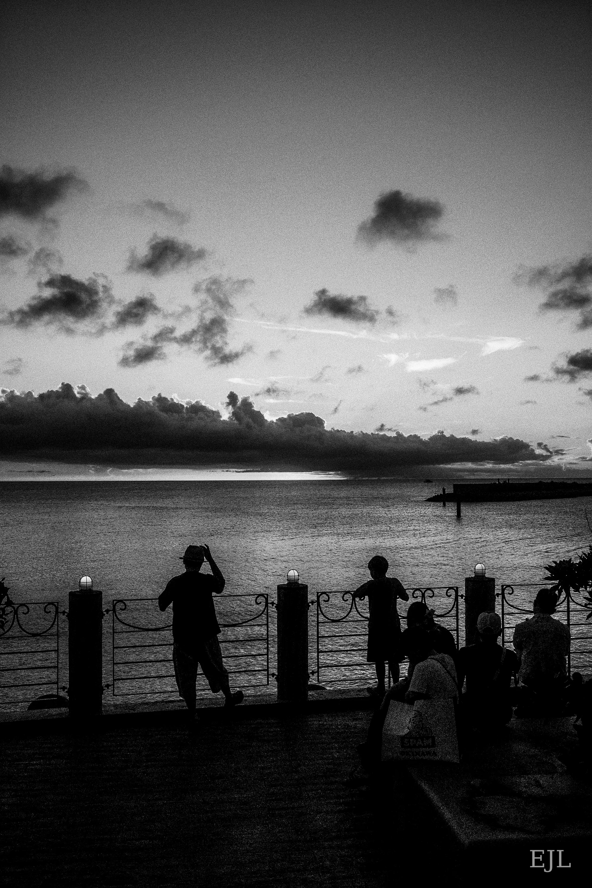 Black-and-white silhouettes beside a waterfront railing beneath a cloud-filled evening sky