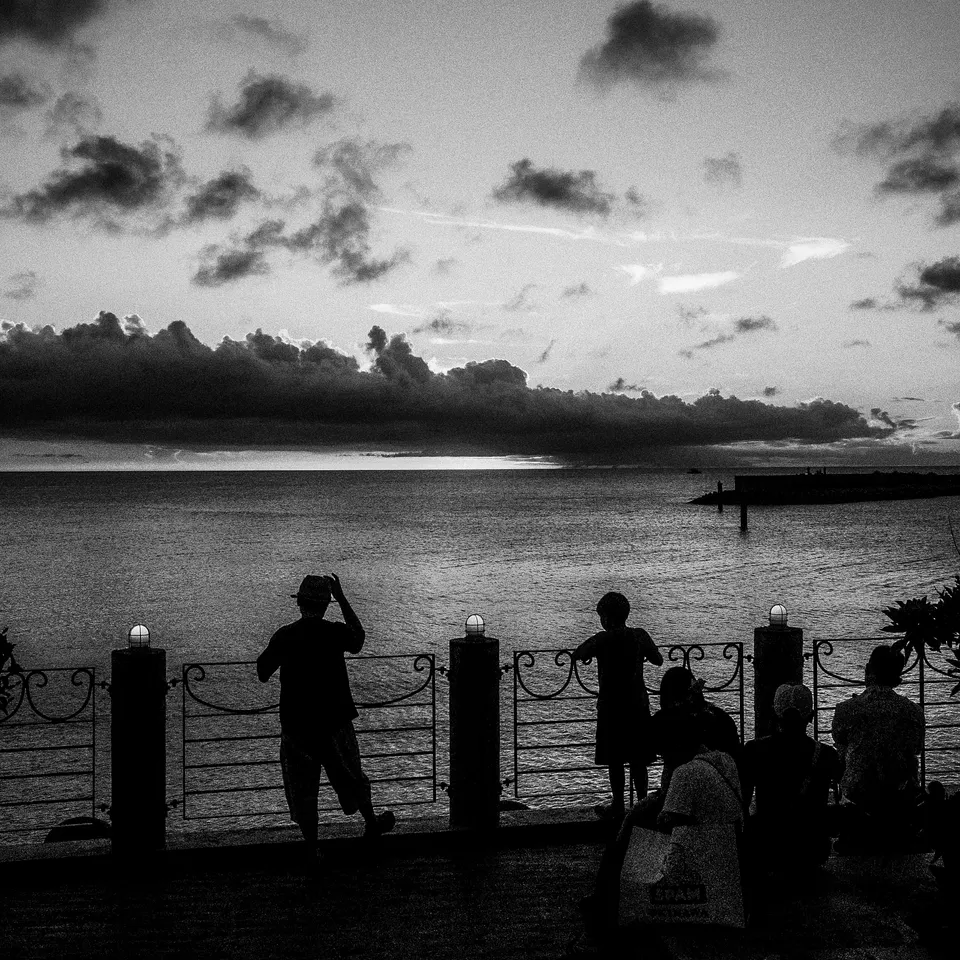 Black-and-white silhouettes beside a waterfront railing beneath a cloud-filled evening sky