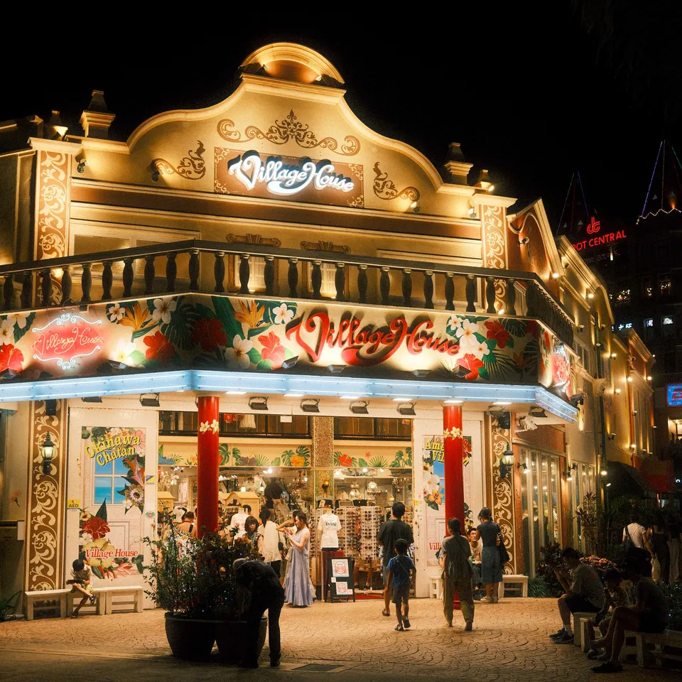 The illuminated Village House storefront with shoppers outside at night