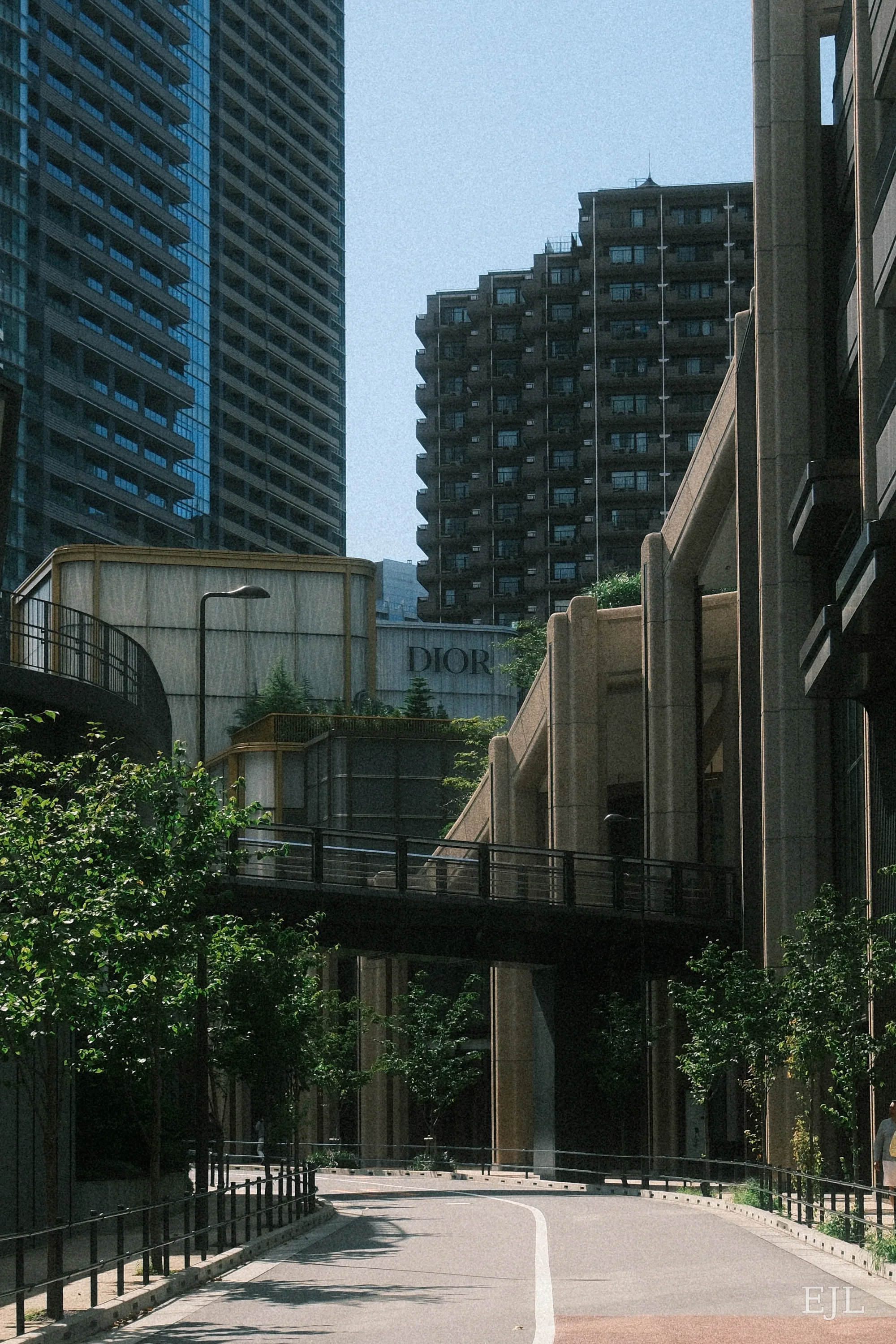 A curving road beneath a pedestrian bridge, surrounded by tall buildings and greenery.