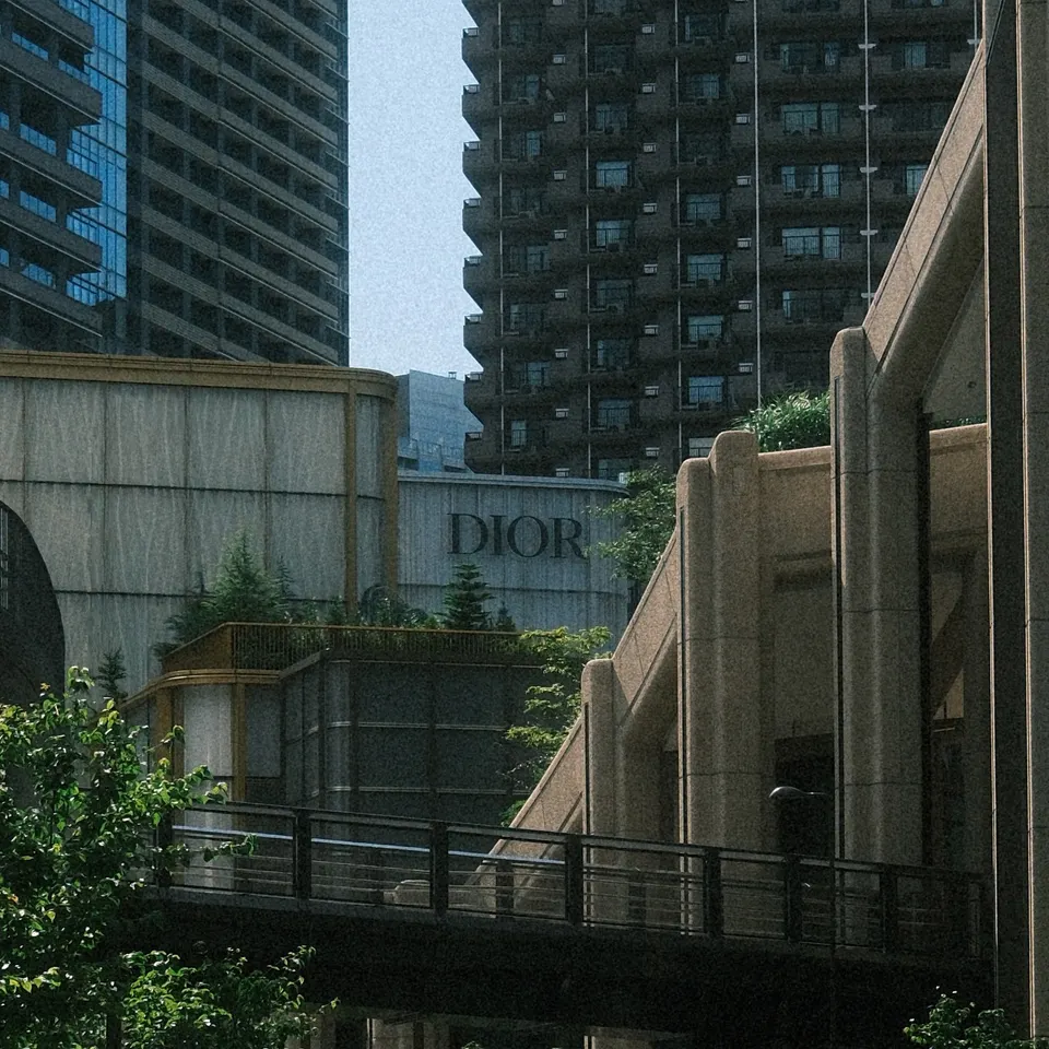 A curving city road beneath a pedestrian bridge, bordered by tall buildings and trees